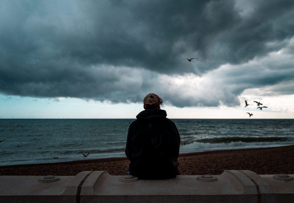 Rear view color image depicting a young man sitting alone at the beach, looking at the dark sea and storm clouds. Moody image emphasising loneliness and isolation. Seagulls are circling and swooping in the distance.